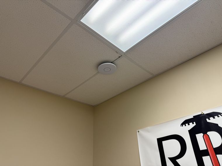 Wireless access point installed on a suspended office ceiling next to a light panel.