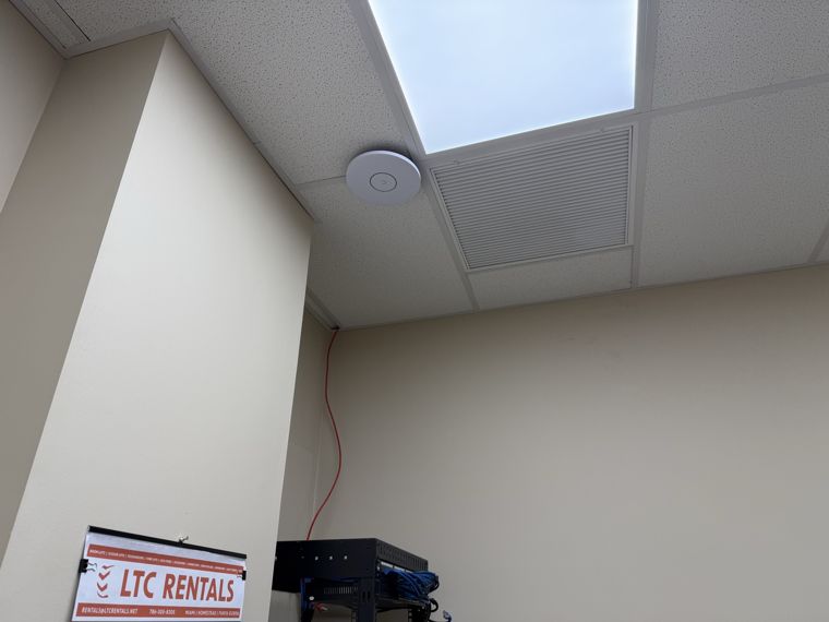 Ceiling-mounted wireless access point beside a light panel and ventilation grille above network equipment.
